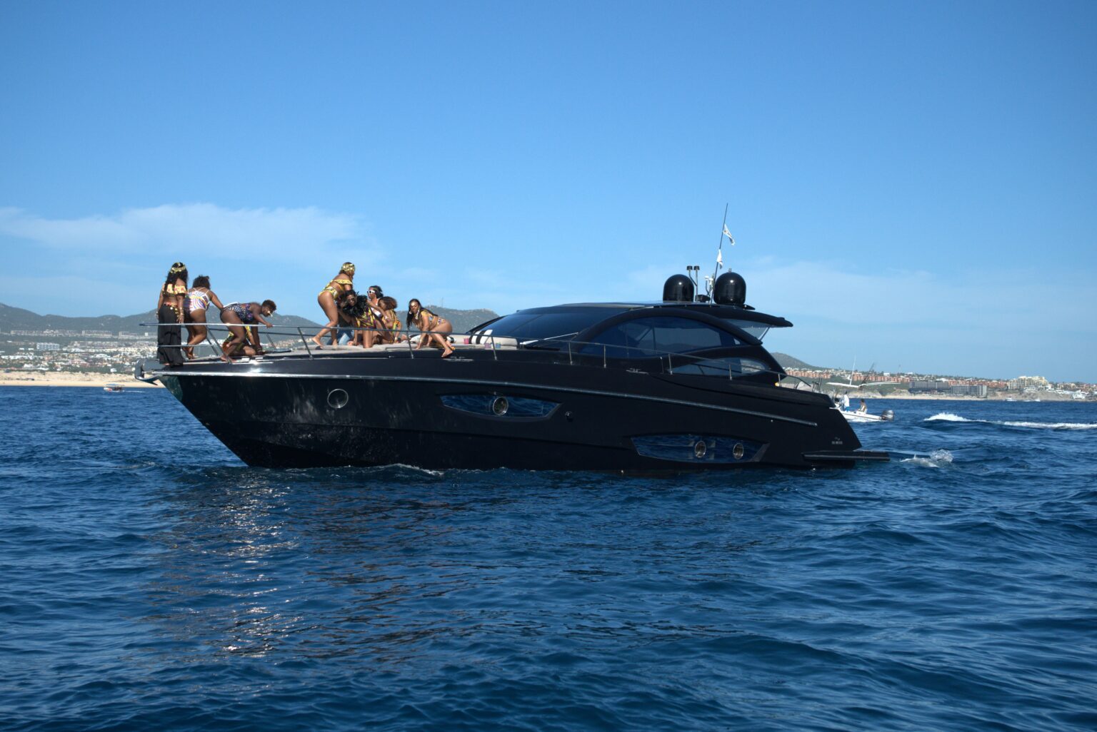 Guests on the bow of a black motor yacht cruising along the coast